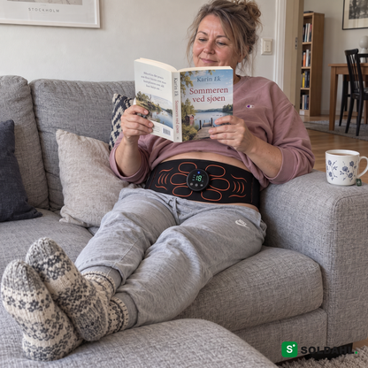 Kvinne leser en bok i sofaen med magebeltet rundt magen