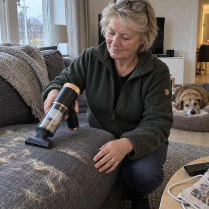 Kvinne fjerner hundehår fra sofaen med Sugro mens hunden hviler i bakgrunnen