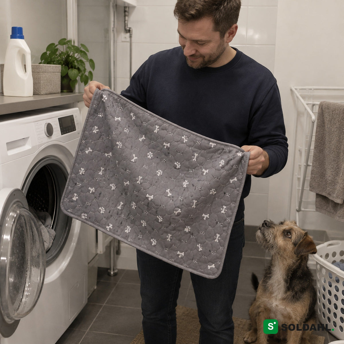 Mann tar den vaskede grå valpematten ut av vaskemaskinen mens hunden ser på